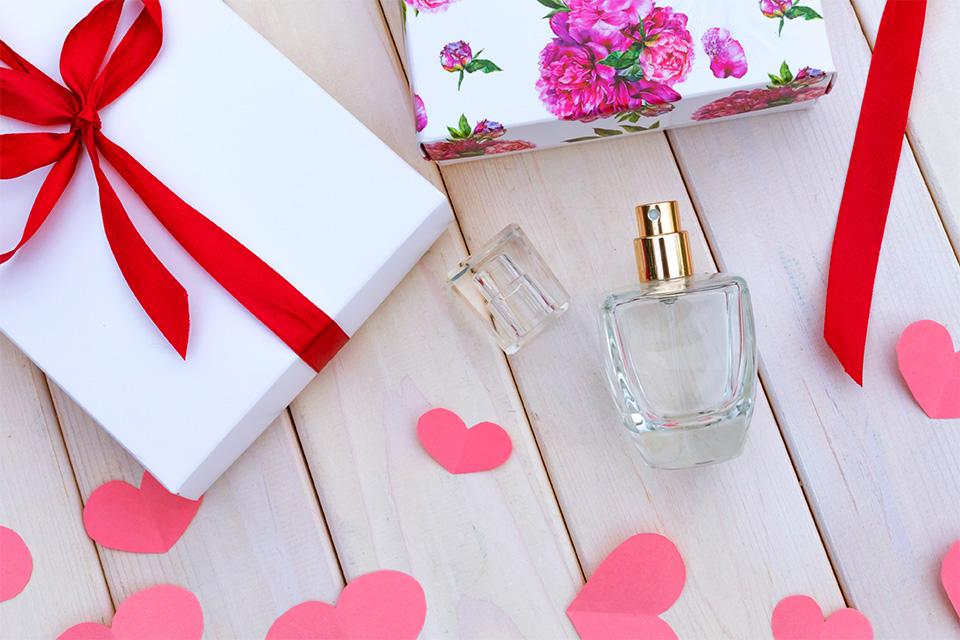 A perfume bottle is next to two presents, wrapped with red ribbon on a wooden floor. Red paper hearts are scattered around below them.