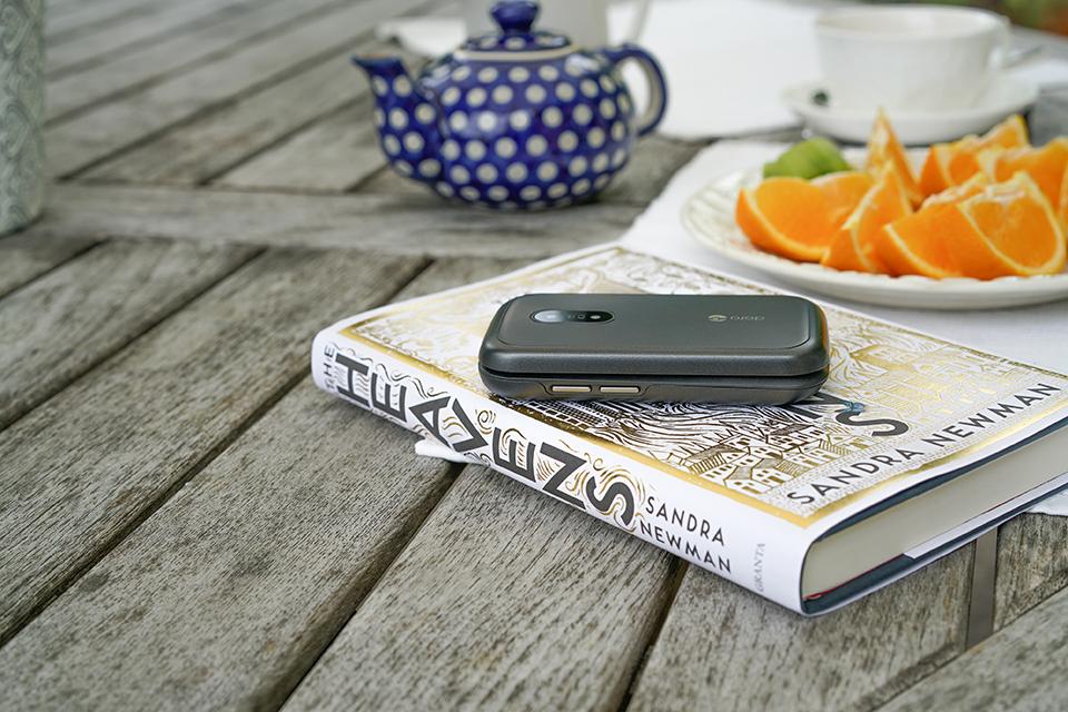 A grey Doro 6040 phone is placed on top of a closed book. Behind is a blue-and-white teapot and a plate of orange slices.