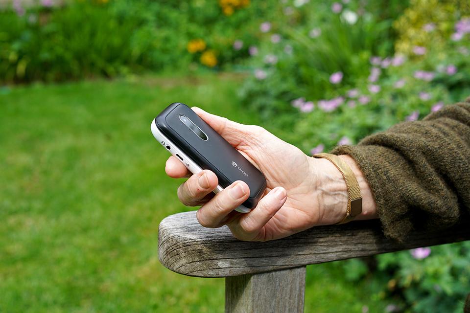 An elderly woman holds a Doro 6620 phone in one hand, resting on the arm of a garden bench.
