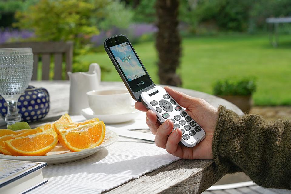 A hand holds a Doro 7030 phone which has been flipped open. The hand is resting on a white table in a sunny garden.