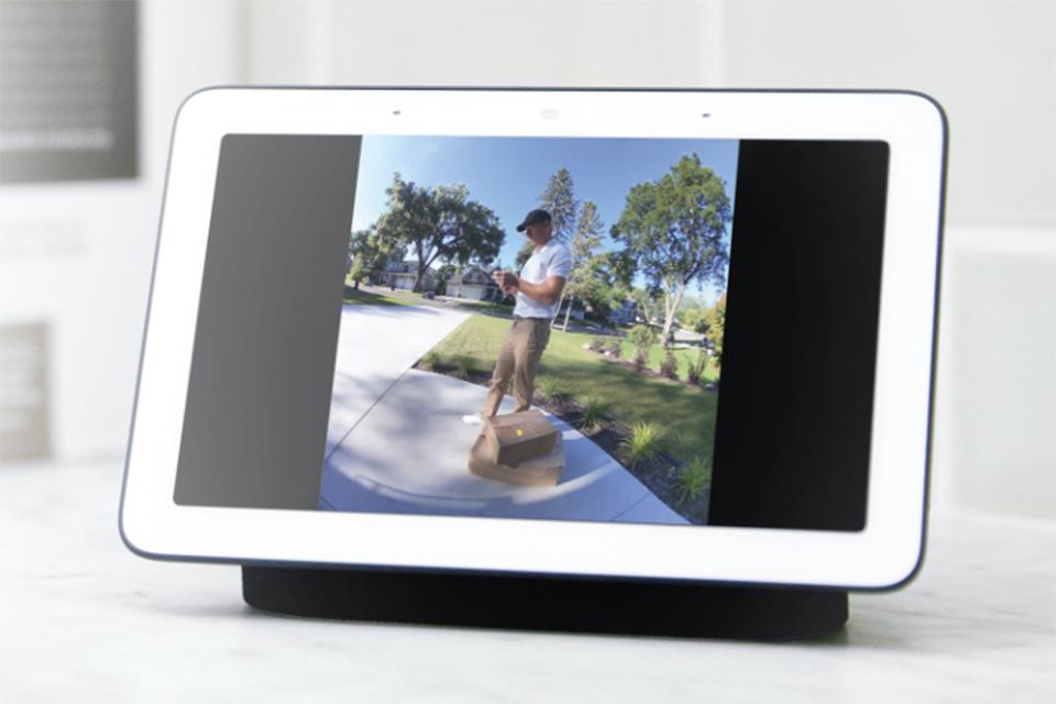 Four smiling men, one holding balloons and one holding a large box, look up towards a fish-eye lens smart doorbell. The image is displayed on a tablet screen.