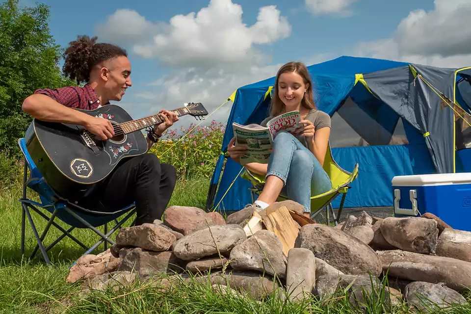 A boy playing his guitar next to a girl reading her magazine outside the tent on a sunny day.