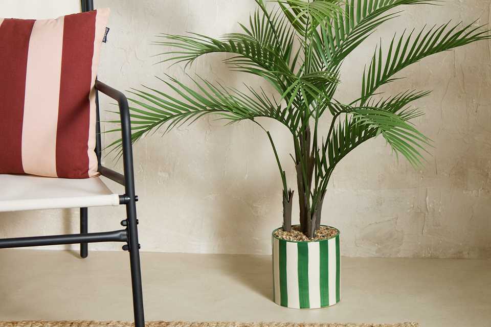 Home 44 inch tall faux plam tree in a green and white striped planter placed next to an armchair.