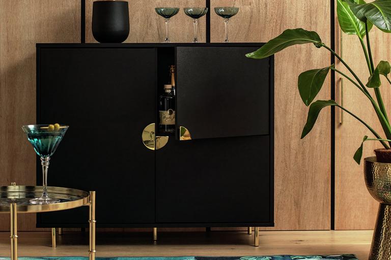 A Habitat 3 door drinks cabinet in black and gold finish with a vase and wine glasses on it and a drinks trolley in front of it.