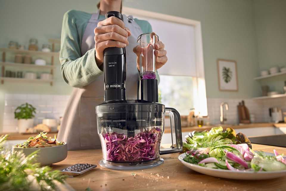 A woman using a Bosch food processor to grate vegetables on a wooden kitchen countertop.