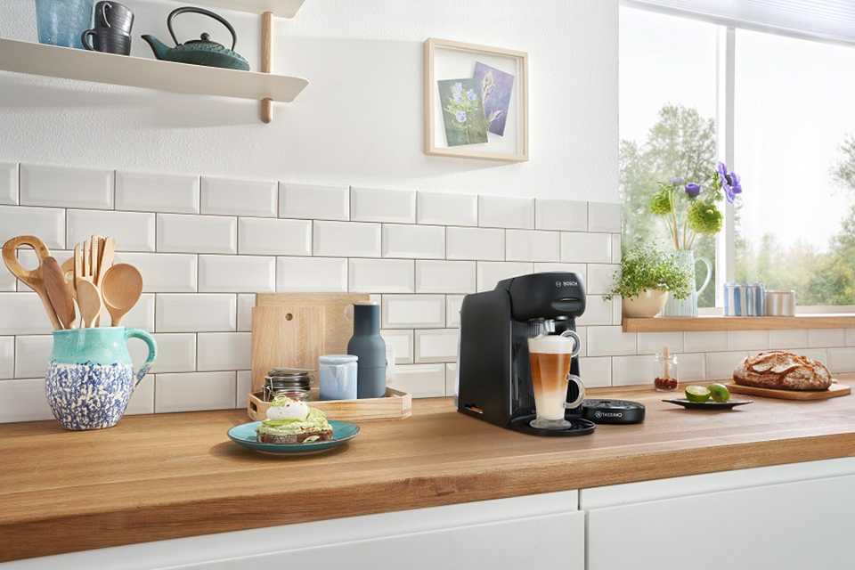A black Tassimo by Bosch pod coffee machine with food items and crockeries on a kitchen countertop.