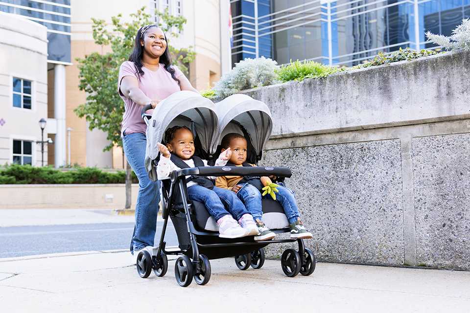 A woman pushing two toddlers in a Graco DuoRider Twin Pushchair - Steeple Gray.