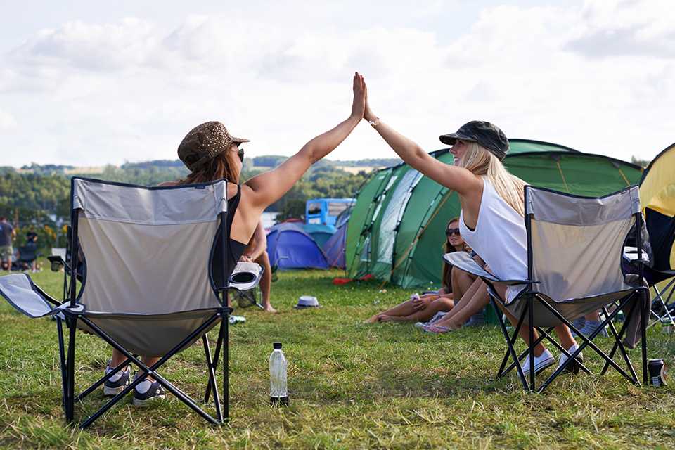 Two friends sitting on camping chairs amidst a festival and giving each other a high five.