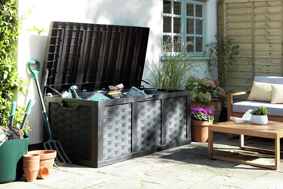 A black McGregor rattan storage box full of gardening tools with plants next to it in a backyard.