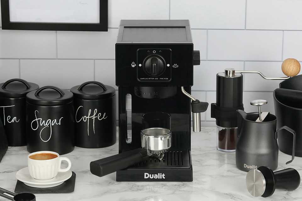 A black Dualit espresso coffee machine with Dualit milk frother, handheld coffee grinder and cups.