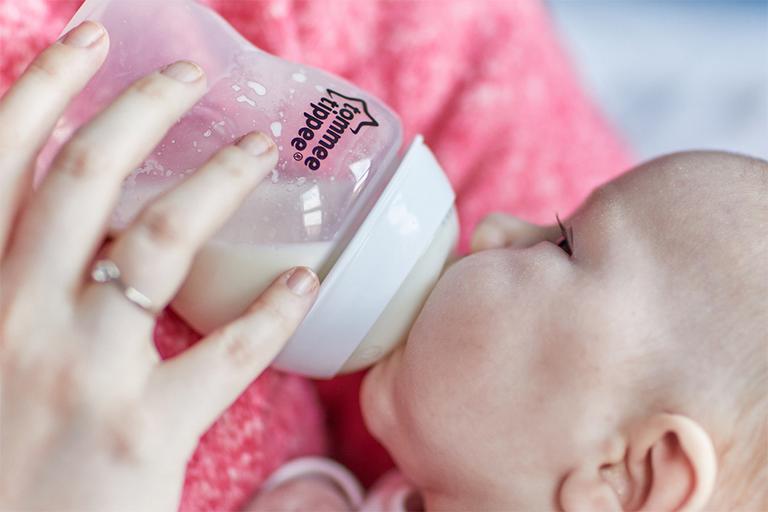 A baby drinks milk from a Tommee Tippee baby bottle. Both baby and bottle are held by someone wearing a pink jumper, with only shoulders visible.