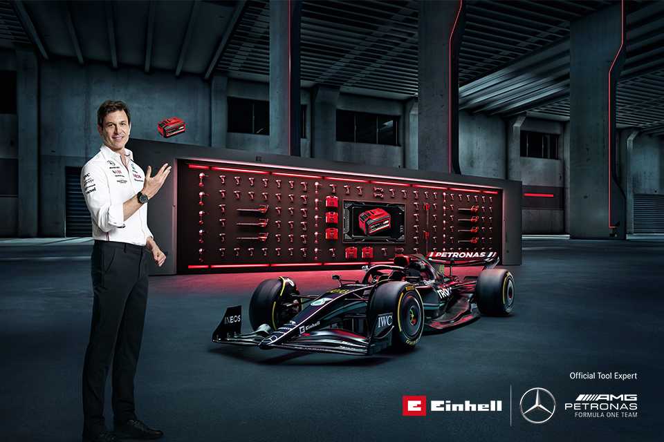 Toto Wolff posing next to a Mercedes Petronas AMG F1 car with Einhell's brand colours and setup in the background and Einhell and Mercedes Petronas AMG logos placed in bottom right corner.