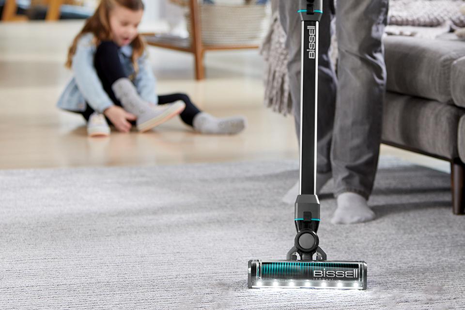 An adult is vacuuming a rug, whilst a child plays in the background.