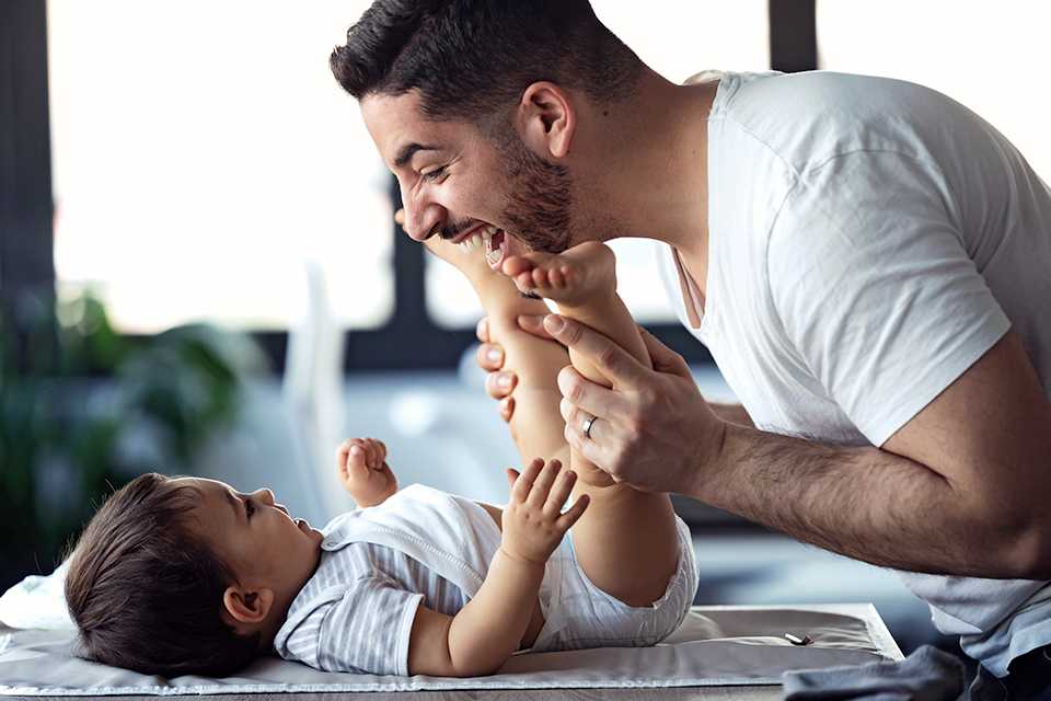 A father playing with a baby while also changing their clothes.