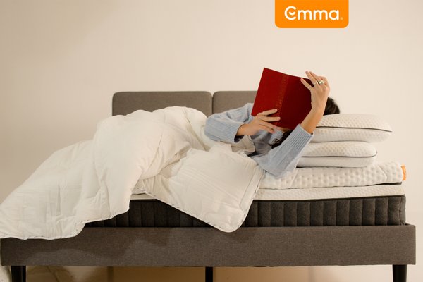Person lying on an Emma mattress with pillows and duvet, reading a book on a bed.