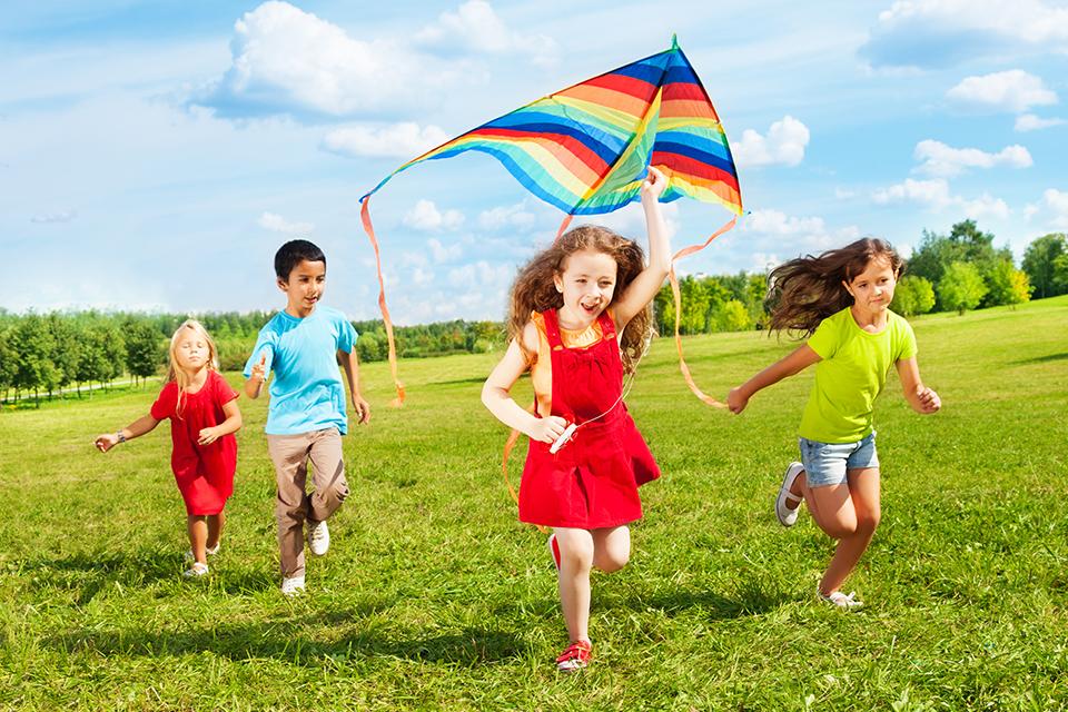 Four children running with one girl trying to launch a colourful kite in the air.