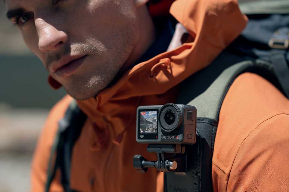 A man wearing an orange coat and carrying a backpack with DJI Osmo Action 4 attached to the strap.