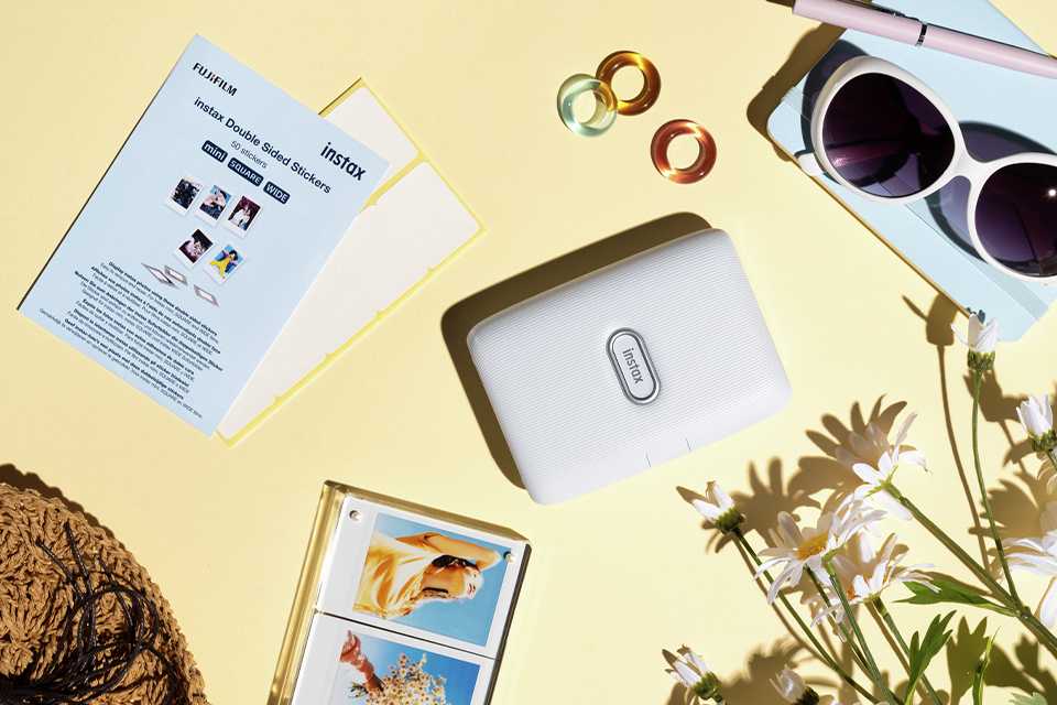 instax Mini Link Smartphone Printer in ash white colour displayed on a yellow background with flowers, prints, and sunglasses placed around it.
