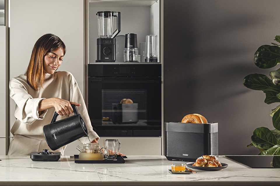 A woman pouring tea from a Haier kettle and toasting some bread using a Haier 2-slot toaster.