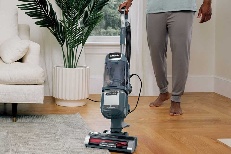 A man using Shark Anti Hair Wrap Plus Pet Upright Corded Vacuum Cleaner in a living room with a dog sitting on a floor nearby and a little girl sitting on the sofa.