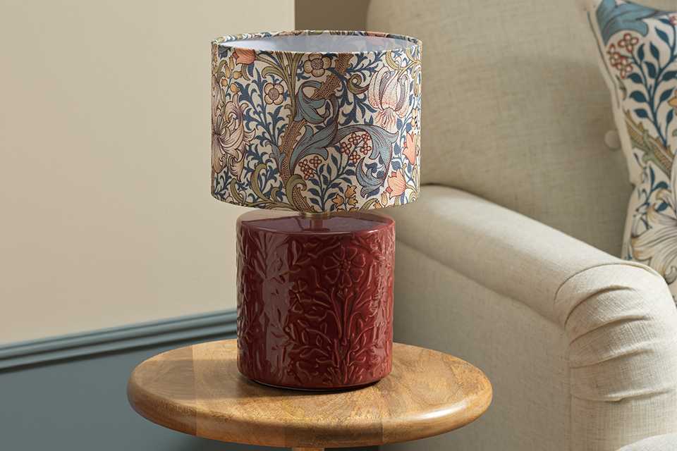 A decorative table lamp with a floral-patterned lampshade and a textured red ceramic base, placed on a wooden side table next to a beige upholstered armchair with a patterned cushion.