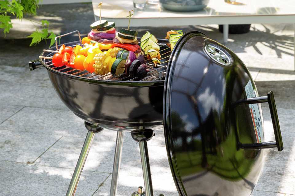 An Argos Home 43cm Kettle Charcoal BBQ grilling vegetables with condiments stored below.
