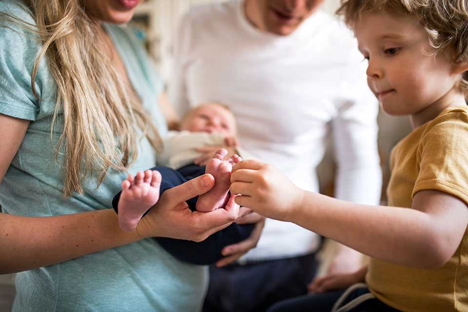 A lady holding a baby while a kid touches their feet with a man in the background.