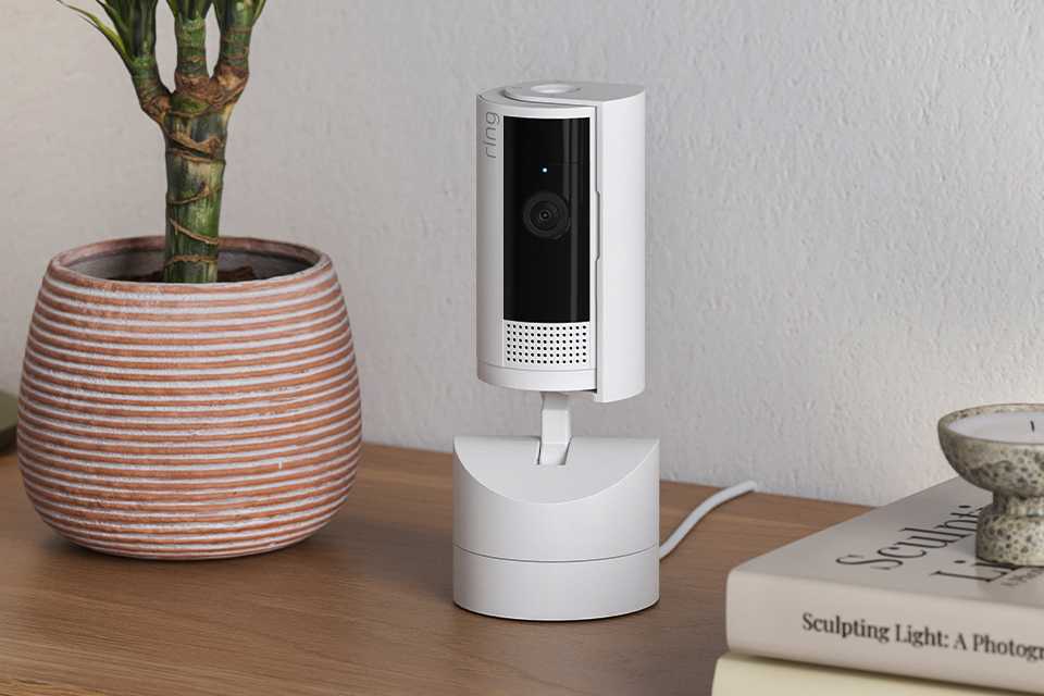 A Ring pan-tilt indoor camera placed on a desk next to a pot of plant.