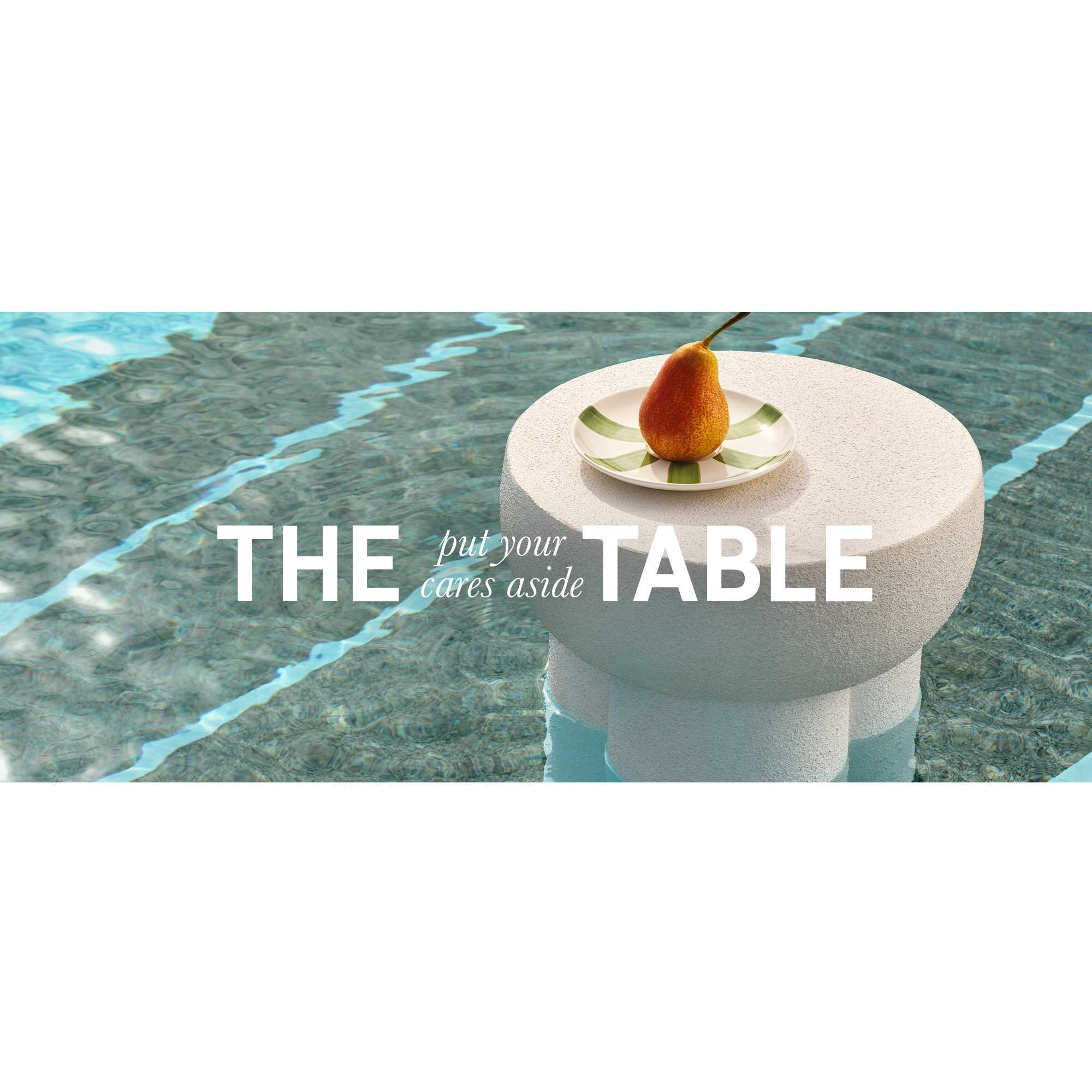 A banner displaying a white Habitat side table in a pool with a pear placed in a plate on top of it.