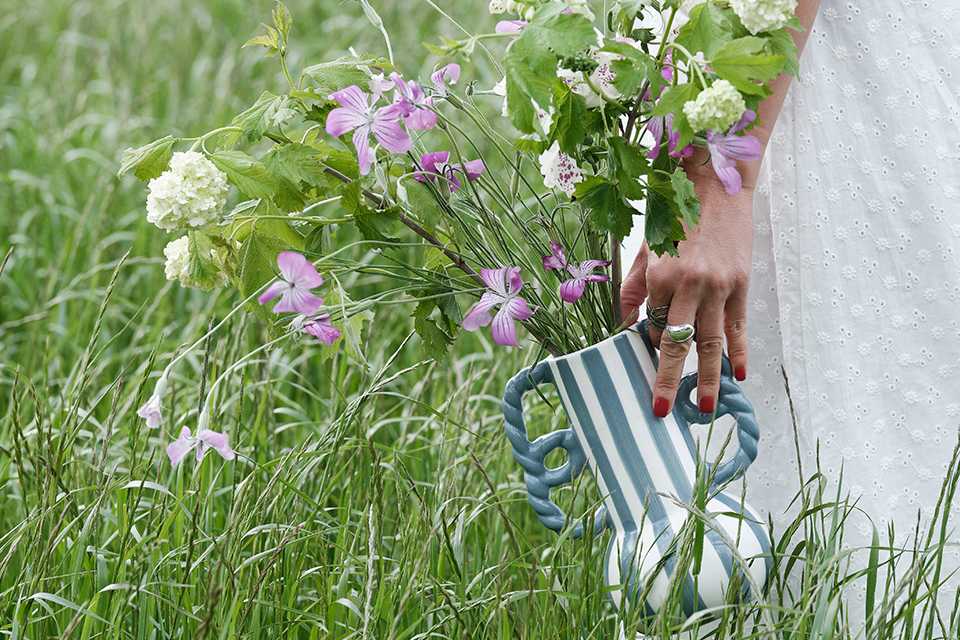A woman holding a Habitat Stripe Ceramic Blue Vase with Wavy Handles and walking outdoors in tall grass.