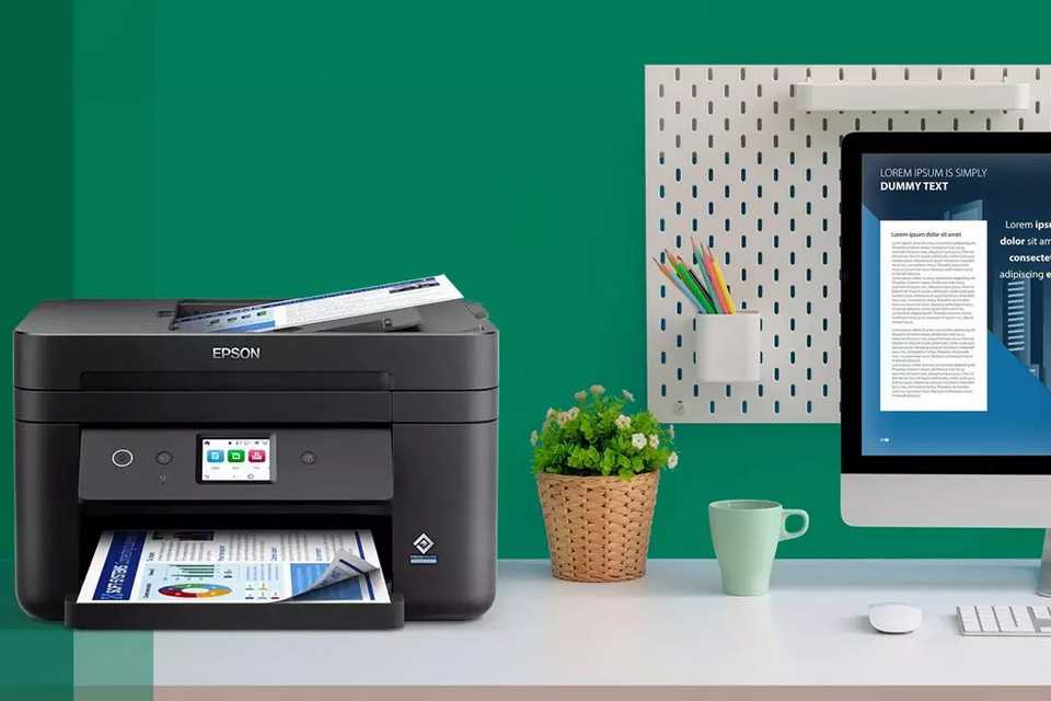 A black Epson printer placed on a work desk against a green wall.