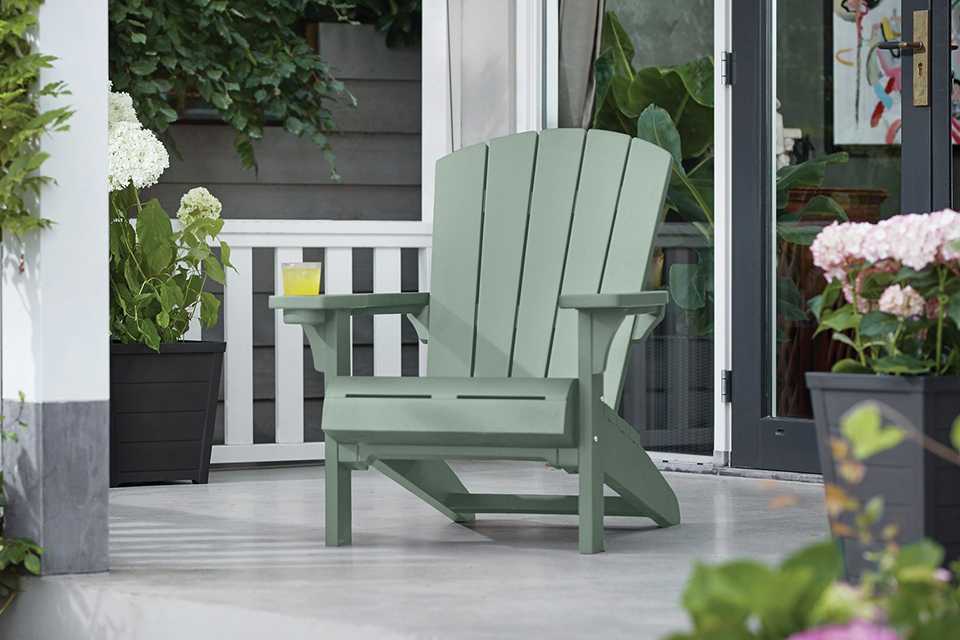 A green Keter Alpine Adirondack resin chair placed outdoors near green plants and flowers.