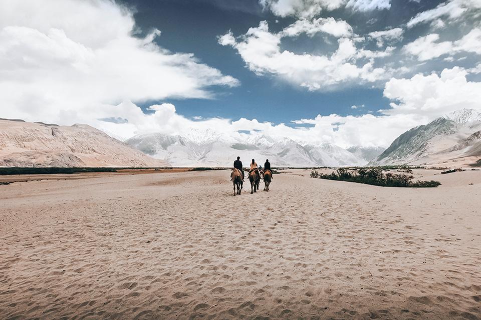 Image shows a panoramic desert landscape, with mountains rising on either side. In the centre, three people ride away from the camera on horseback.