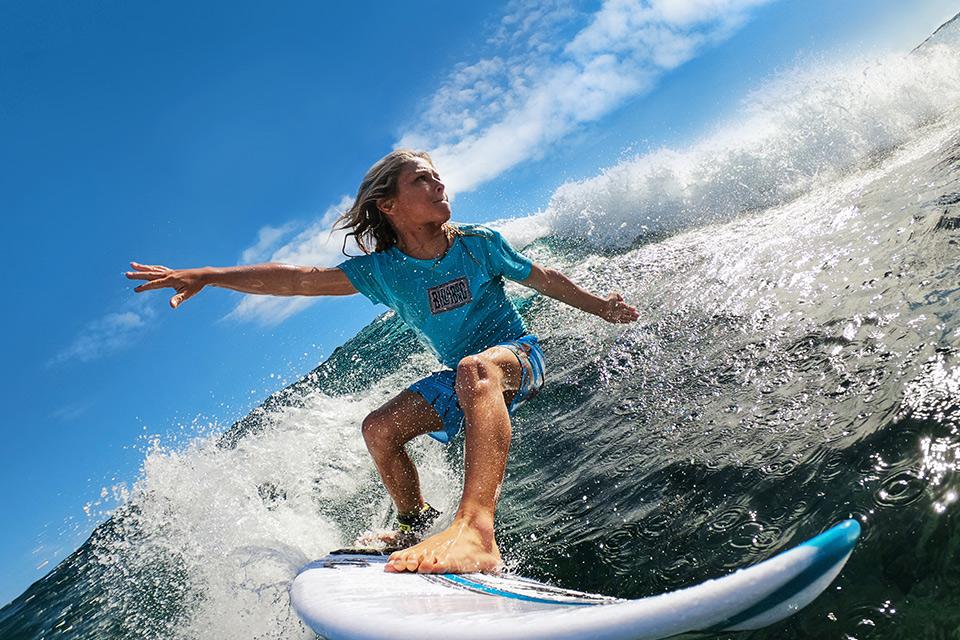 A surfer rides a tall wave. The low-angle shot captures the moment the wave curves and crashes into foam, and blue sunny skies.