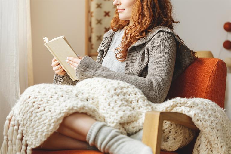 A woman sat cosily reading in a chair with a blanket on her lap.