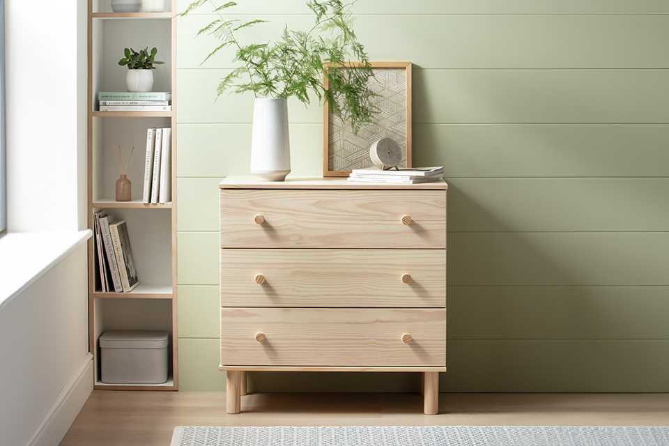 A wooden Habitat 3 drawer chest in a bedroom with a picture frame and a table lamp on top of it.