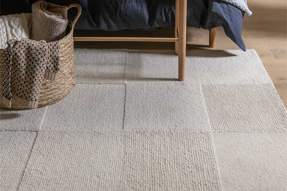 Habitat Checkerboard rug in neutral tones, placed in a bright, modern living room. The wool rug features a bold high-low checkerboard pattern with a soft cut pile texture, adding warmth and visual interest to the wooden floor.