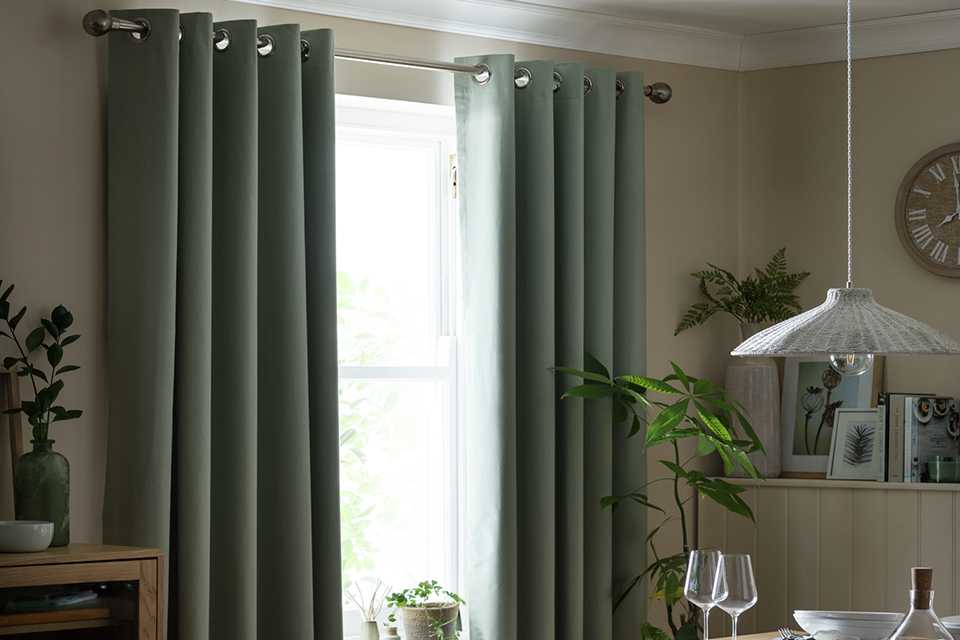 Stylishly hung sage green blackout eyelet curtains from Habitat, displayed against a white wall with natural light filtering through. The curtains feature a smooth, plain texture and silver metal eyelets, offering a modern and minimalist look.