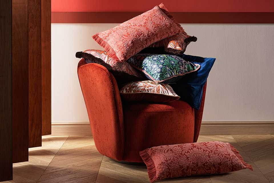 A red cushioned chair adorned with colorful patterned throw pillows in shades of red, blue, green, and orange, with one pillow placed on the floor, set against a two-tone wall and light wood flooring.