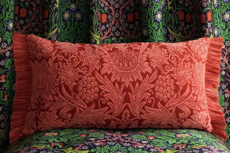 Red floral print long cushion with tuffed edge infront of green floral curtains.