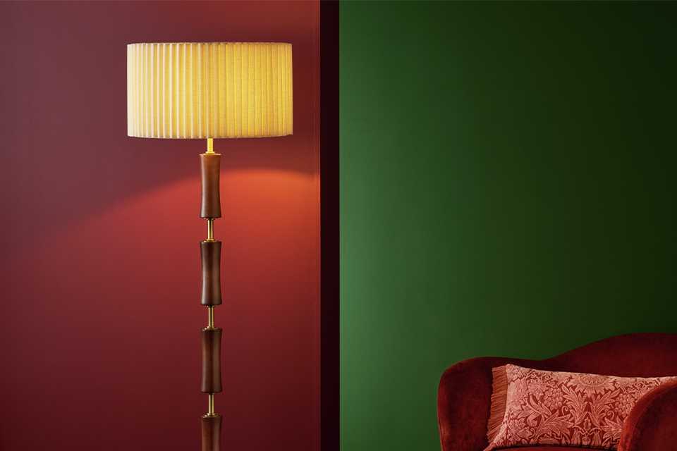 Wooden floor lamp with white shade in ribbed style with orange velvet chair in background infront of green and red walls.