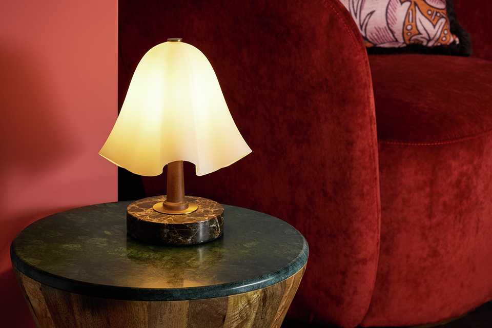 Small short mushroom glas lamp with marble base on side table in front of red velvet chair.