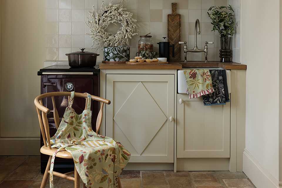 Cosy kitchen corner with a floral apron on a wooden chair, cream cabinet, wooden countertop, and light tiled backsplash.