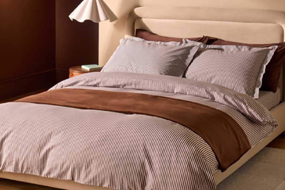 Cream bed in bedroom with stripe brown bedding and wavy white ceiling light.