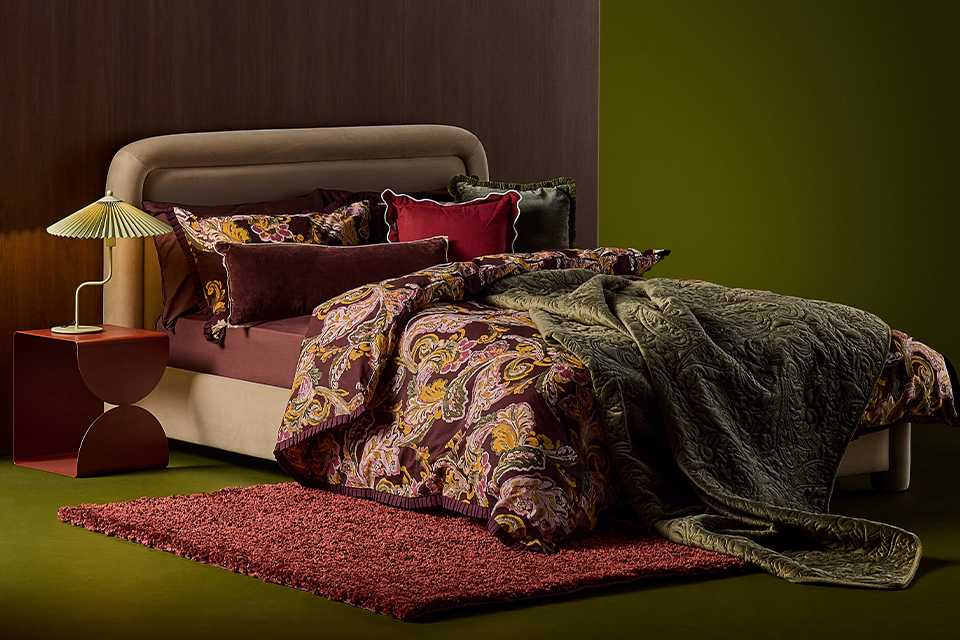 A cosy bed with pillows, a duvet and a throw with a rug under it and a table lamp on a bedside table.