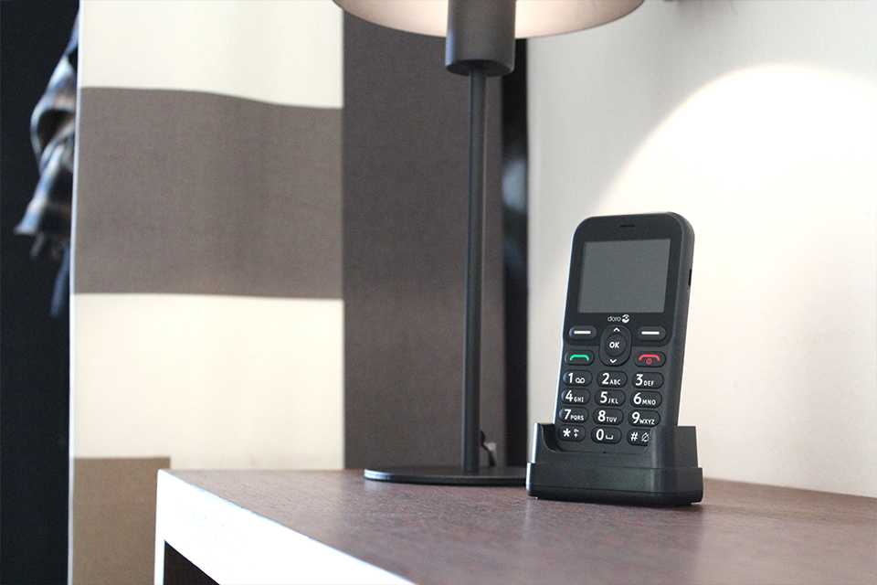 A black Doro Leva E10 feature phone placed in a charging cradle on a sideboard.