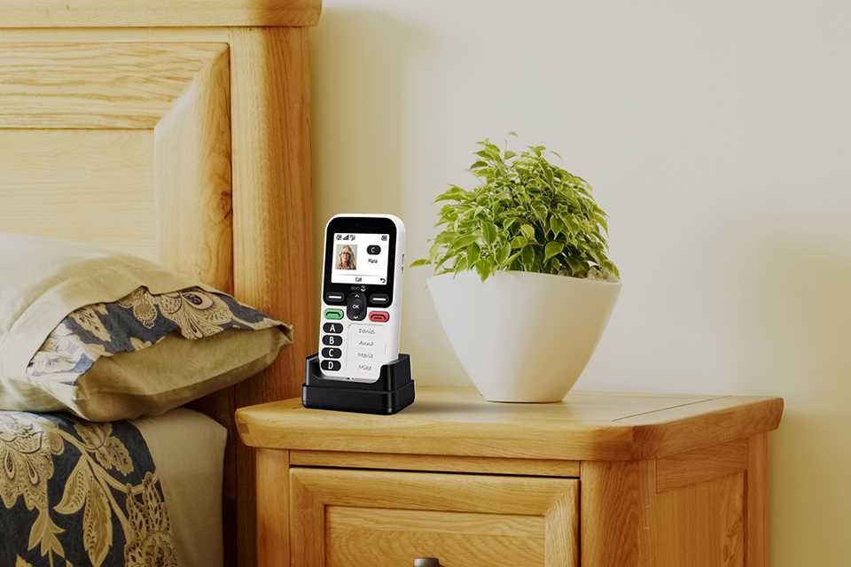 Doro Leva X10 feature phone placed in a charging cradle on a bedside table next to a potted plant.