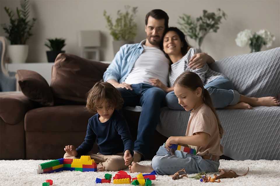 An icon depicting tAn family of 4 sitting in a living room with parents on the sofa and kids playing with blocks on the floor. wo kids playing and dancing.