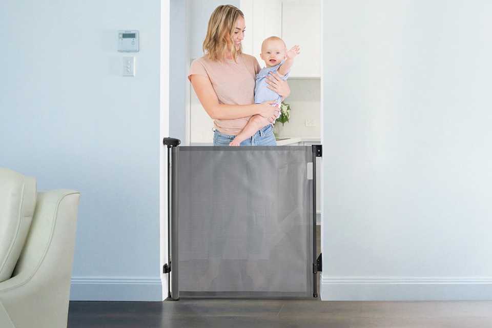 A mother holding her baby and standing behind a Dreambaby X-Tall Retractable Gate. Fits 60-140cm - Grey.