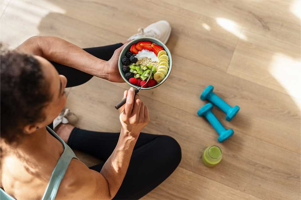 A woman having breakfast sitting on the floor with blue dumbbells and a bottle of healthy juice placed next to her.